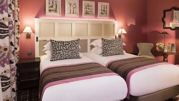 A hotel room with two beds, a headboard, wall-mounted lamps, a chair, a small table with a vase, and a patterned curtain.