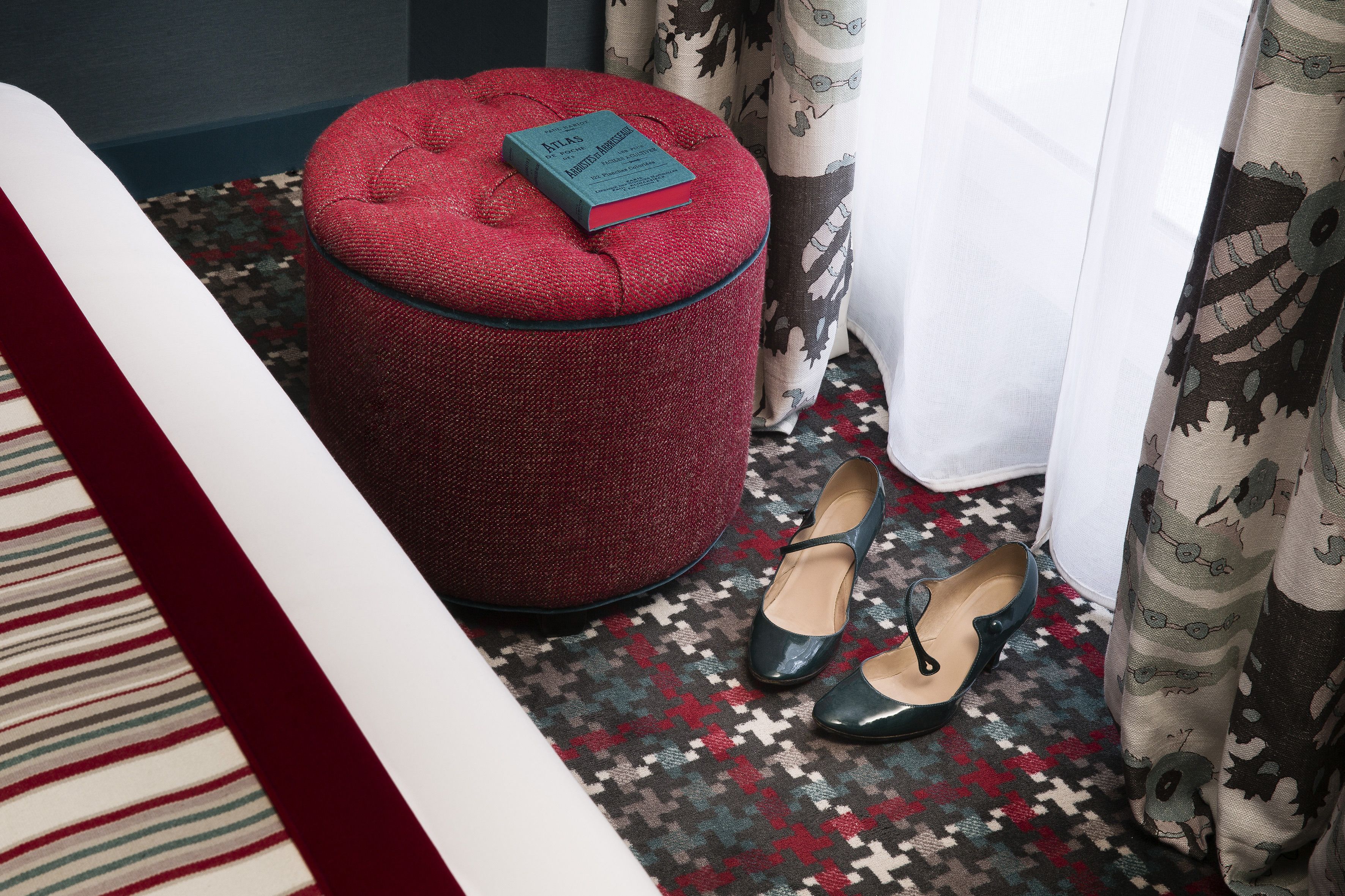 A red ottoman with a book on top, a pair of green shoes, and a patterned carpet in a room with a bed and curtains.