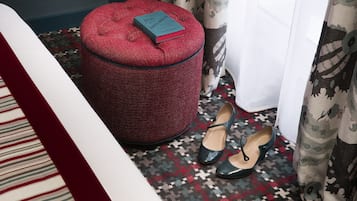 Un reposapiés rojo con un libro encima, un par de zapatos verdes y una alfombra estampada en una habitación con cama y cortinas.