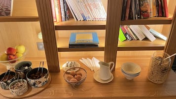 Una mesa de madera con un cuenco de frutas, una jarra y un tarro de granola. Detrás de la mesa, hay una estantería con varios libros y un libro azul titulado