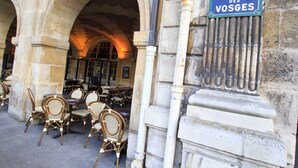 An outdoor seating area with wicker chairs and tables, stone arches, and a sign reading "Place des Vosges". - Les Tournelles (Paris)