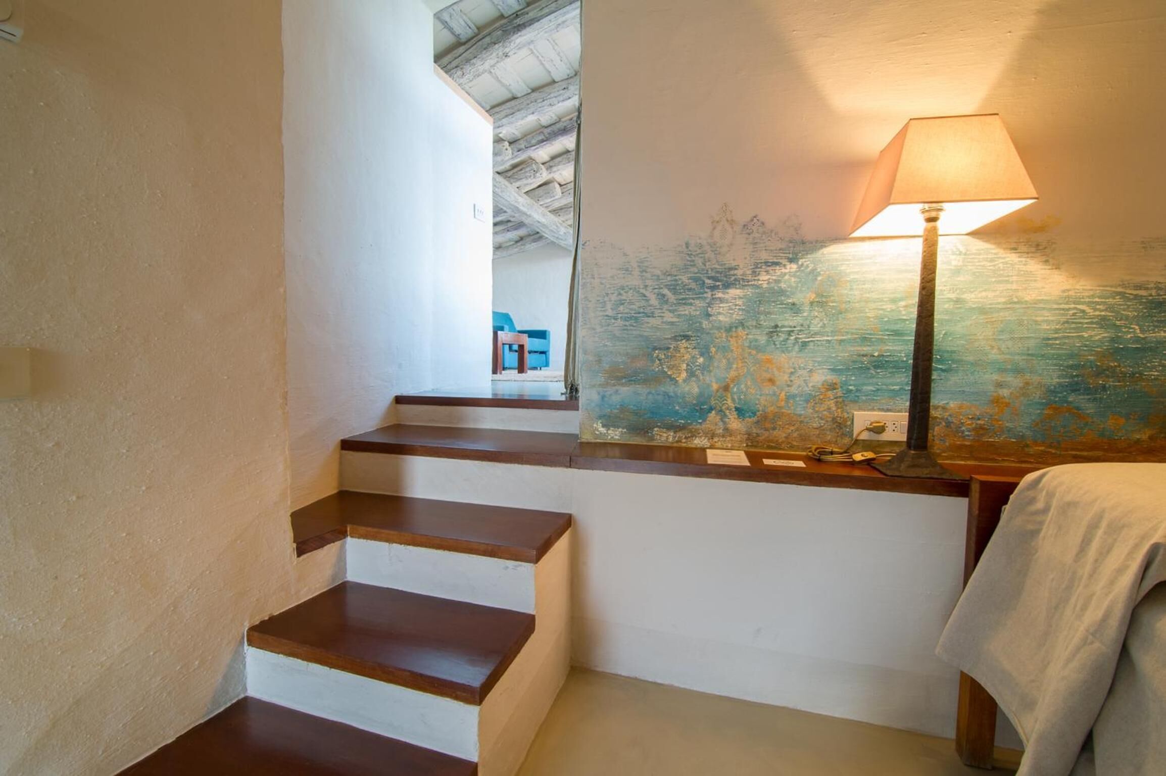 Una escalera de madera con peldaños que conducen a una plataforma, una pared con una pintura texturizada y una mesa con una lámpara.