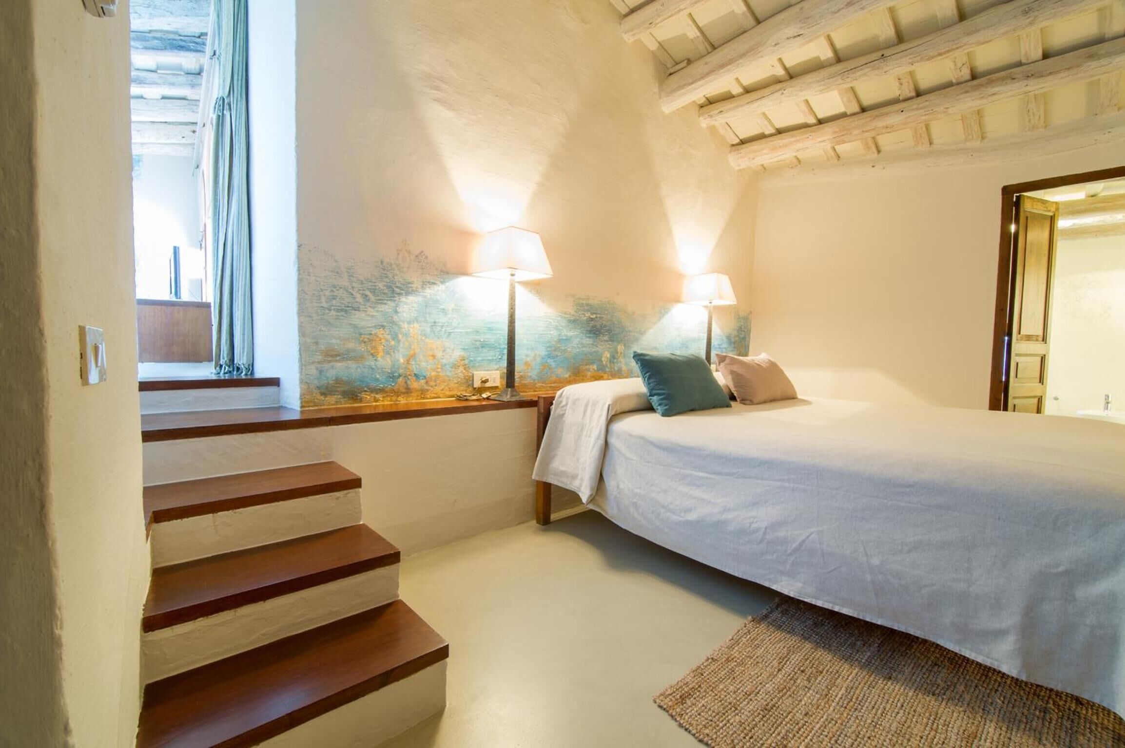 Un dormitorio con cama, dos lámparas de noche, una ventana con cortinas, una escalera de madera y una pared con textura.