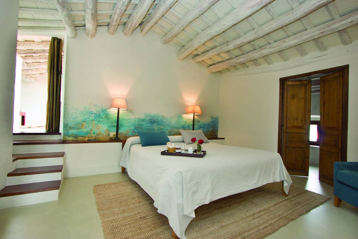 Une chambre à coucher avec un lit, deux lampes de chevet, un tableau au mur, un plateau avec divers objets sur le lit, une chaise bleue et une porte en bois.
