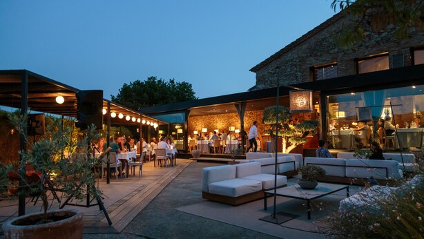 Un espace extérieur aménagé pour les repas, avec des tables et des chaises, un coin salon avec des canapés, et un bâtiment en pierre en arrière-plan.