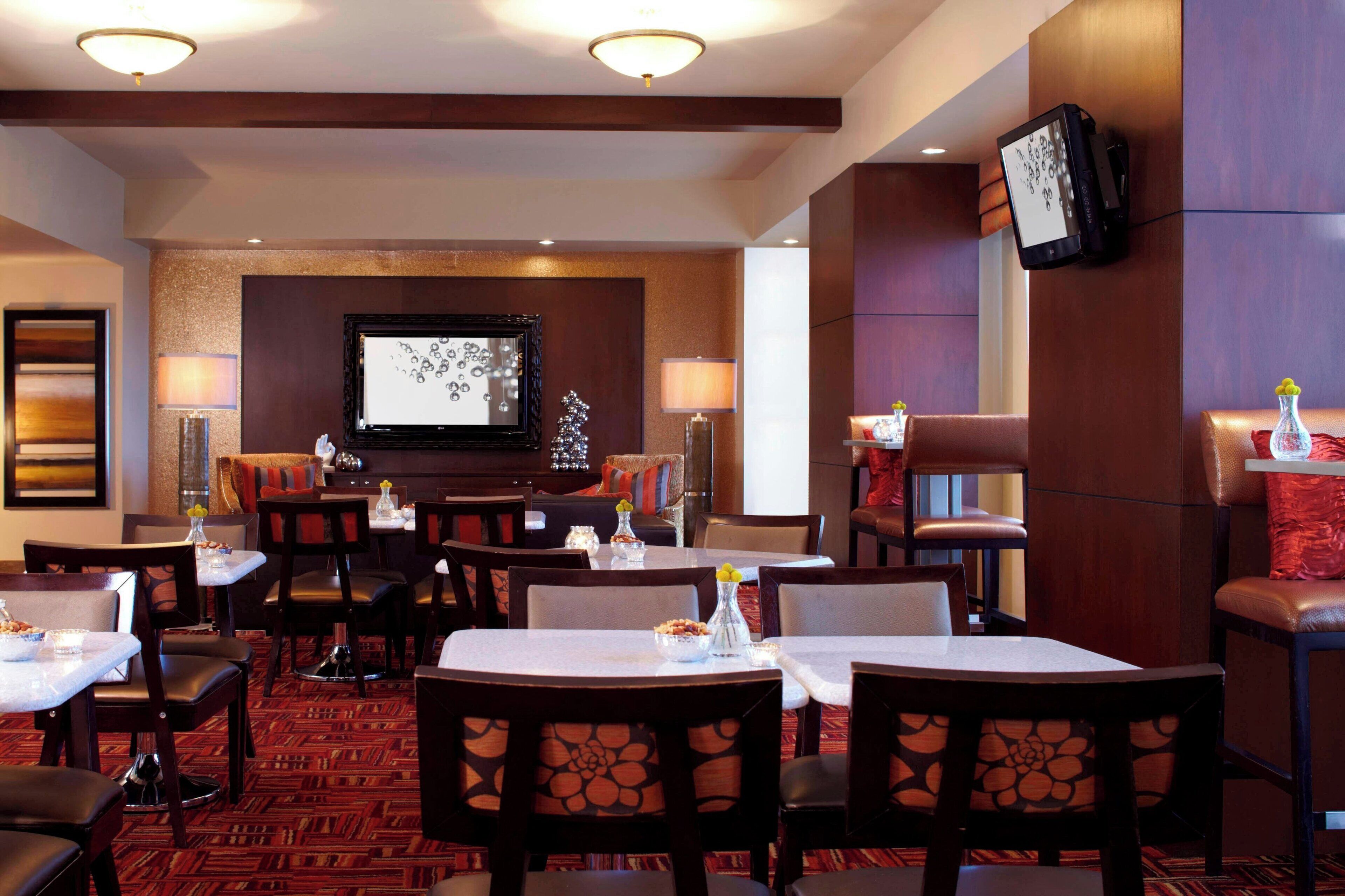 Uma sala de jantar de hotel com várias mesas e cadeiras, uma televisão fixada na parede e um tapete estampado.