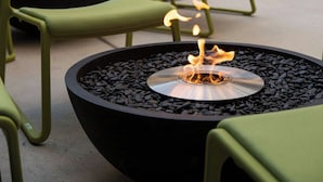 A circular fire pit with a central flame, surrounded by green chairs in an outdoor seating area with a modern building in the background.