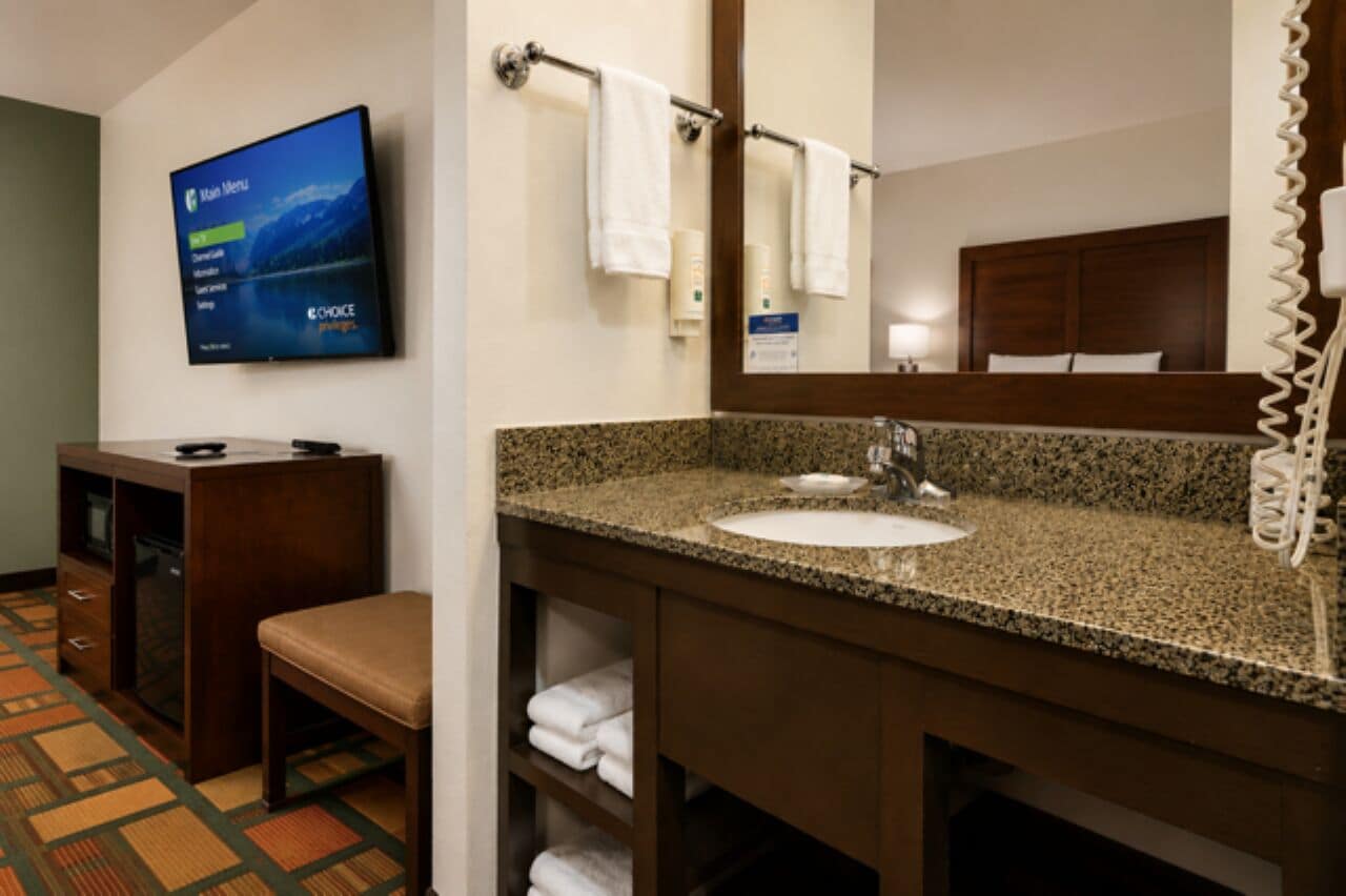 A hotel bathroom with a granite countertop sink, a mirror, a towel rack, a television displaying a menu, and a telephone.