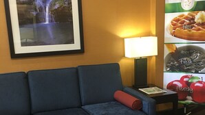 Habitación de hotel con sofá azul, una mesita con lámpara, un cuadro de una cascada y un cartel publicitario con opciones de desayuno.