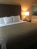 Camera d'albergo con un letto grande, cuscini bianchi, un comodino con una lampada e uno specchio.