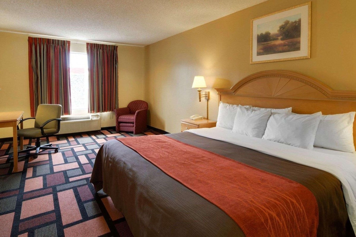 A hotel room with a large bed, a desk with a chair, a window with curtains, and a painting on the wall.
