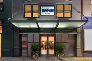 Exterior do Hotel SoLita SoHo com uma placa grande, portas de vidro e duas plantas em vasos na entrada.
