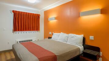 A hotel room with a bed, a nightstand, a wall-mounted lamp, and a window with orange curtains.
