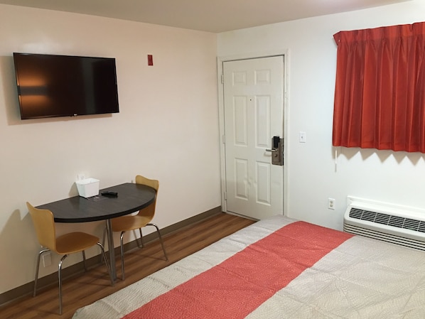 A small, clean room with a flat-screen TV mounted on the wall, a white door with a handle, a red curtain, a white radiator, and a small table with two chairs.