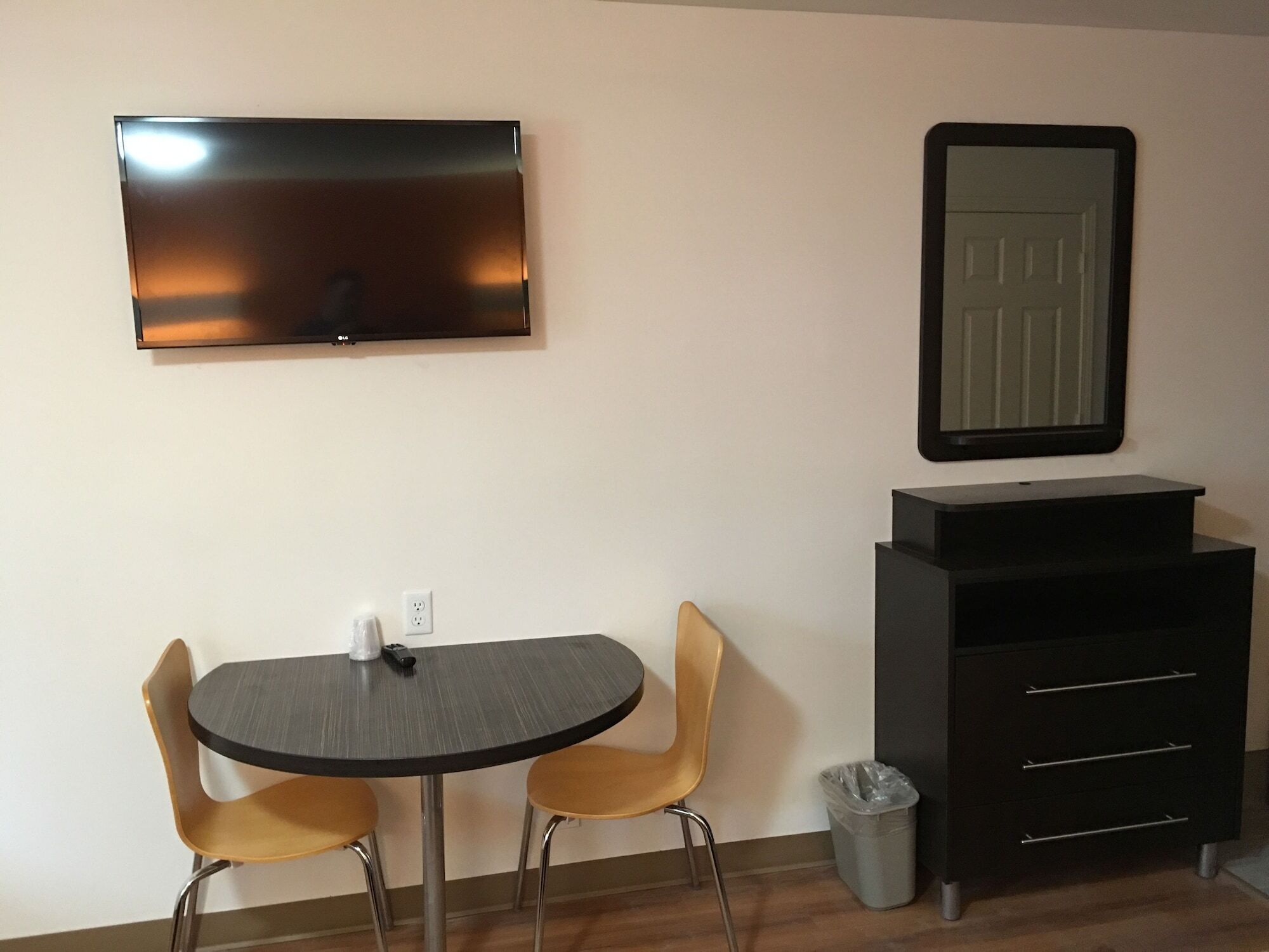 A small room with a round table, two chairs, a TV mounted on the wall, a mirror above a black cabinet, and a trash bin.