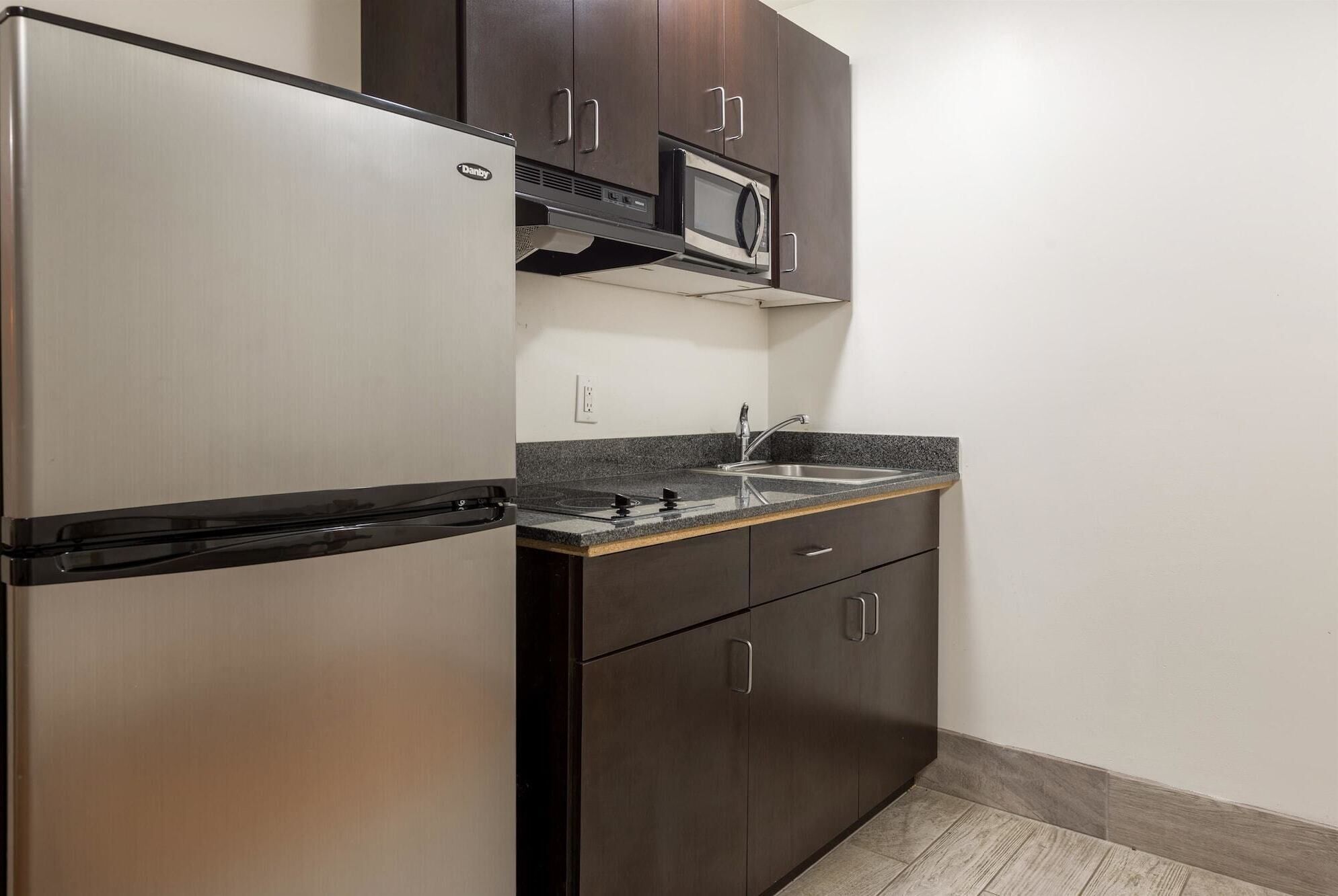 A compact kitchen with a stainless steel refrigerator, a microwave, and a sink with a granite countertop.