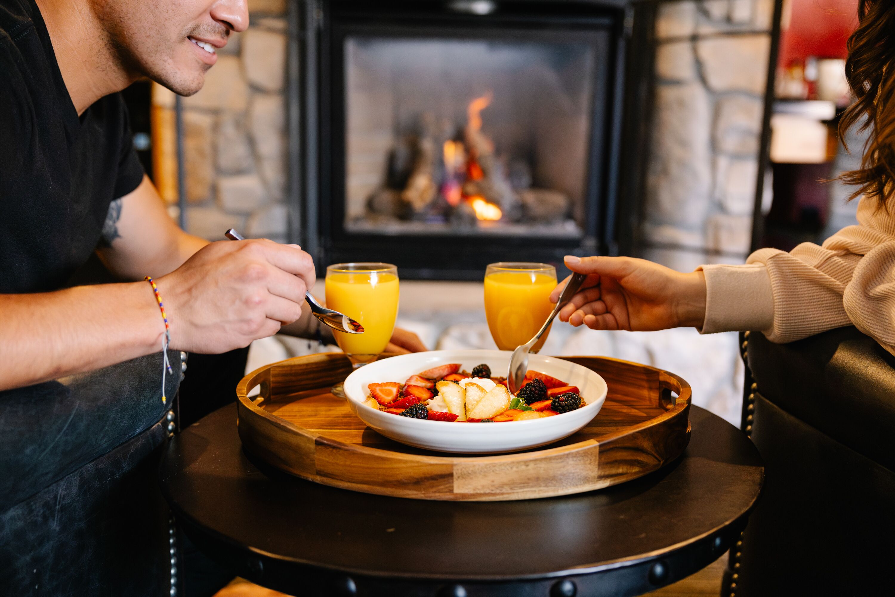 Una pareja cenando en una mesa con una bandeja de madera, un plato de fruta y dos vasos de jugo de naranja. Se ve una chimenea al fondo.