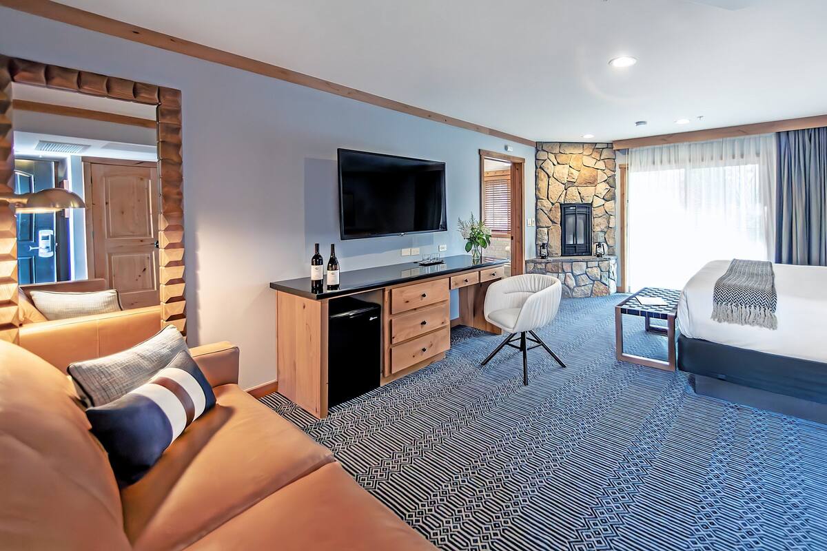 A hotel room with a bed, a desk with a TV, a seating area, and a stone fireplace.