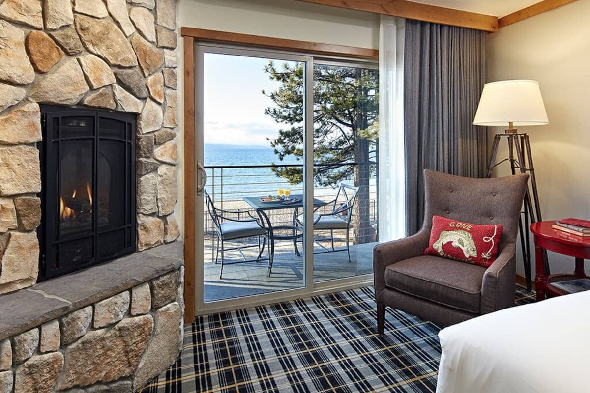 A hotel room with a stone fireplace, a view of the ocean, a balcony with outdoor furniture, and a plaid carpet.