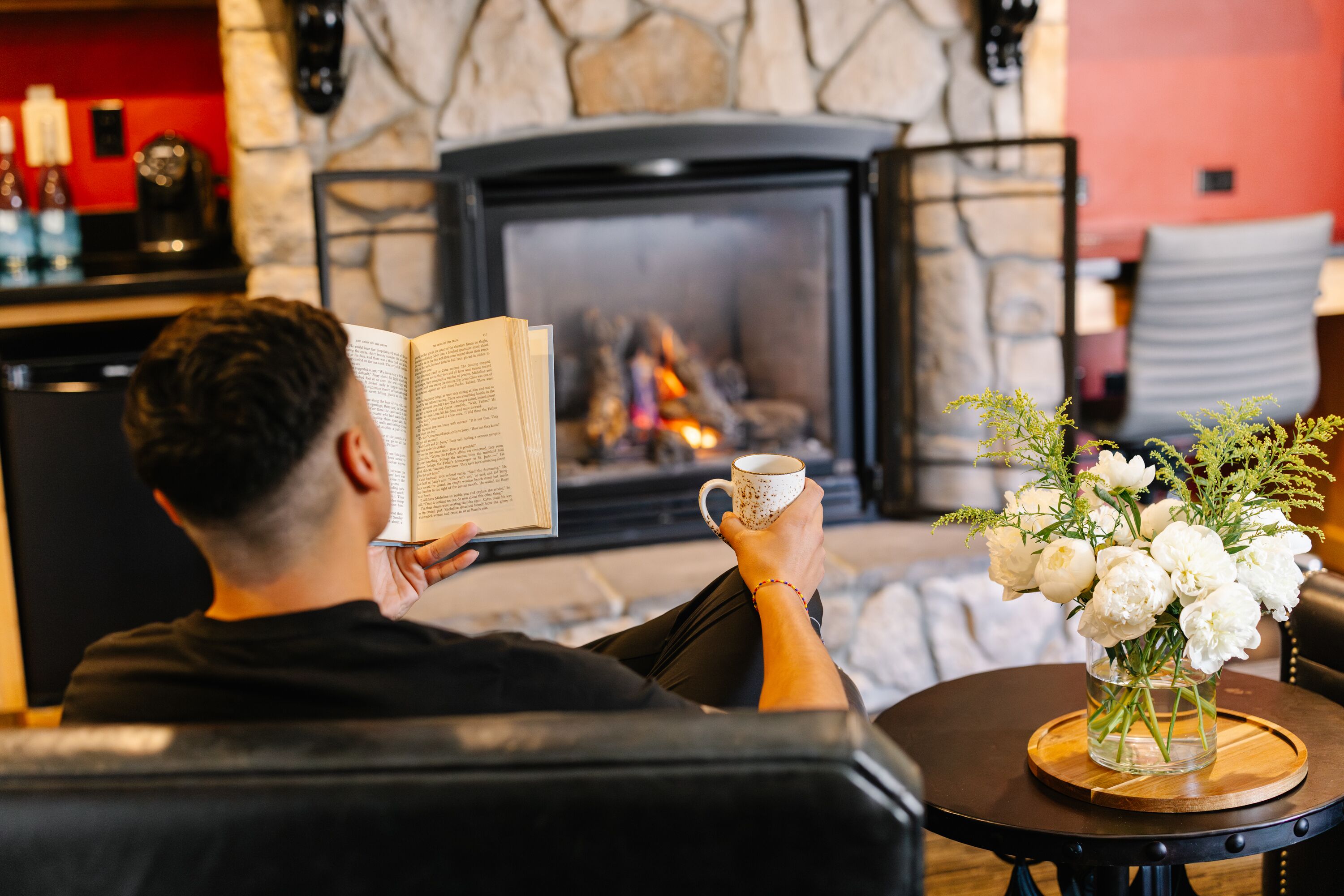 A person reading a book by a fireplace with a mug and flowers on a table.
