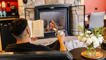 A person reading a book by a fireplace with a mug and flowers on a table.