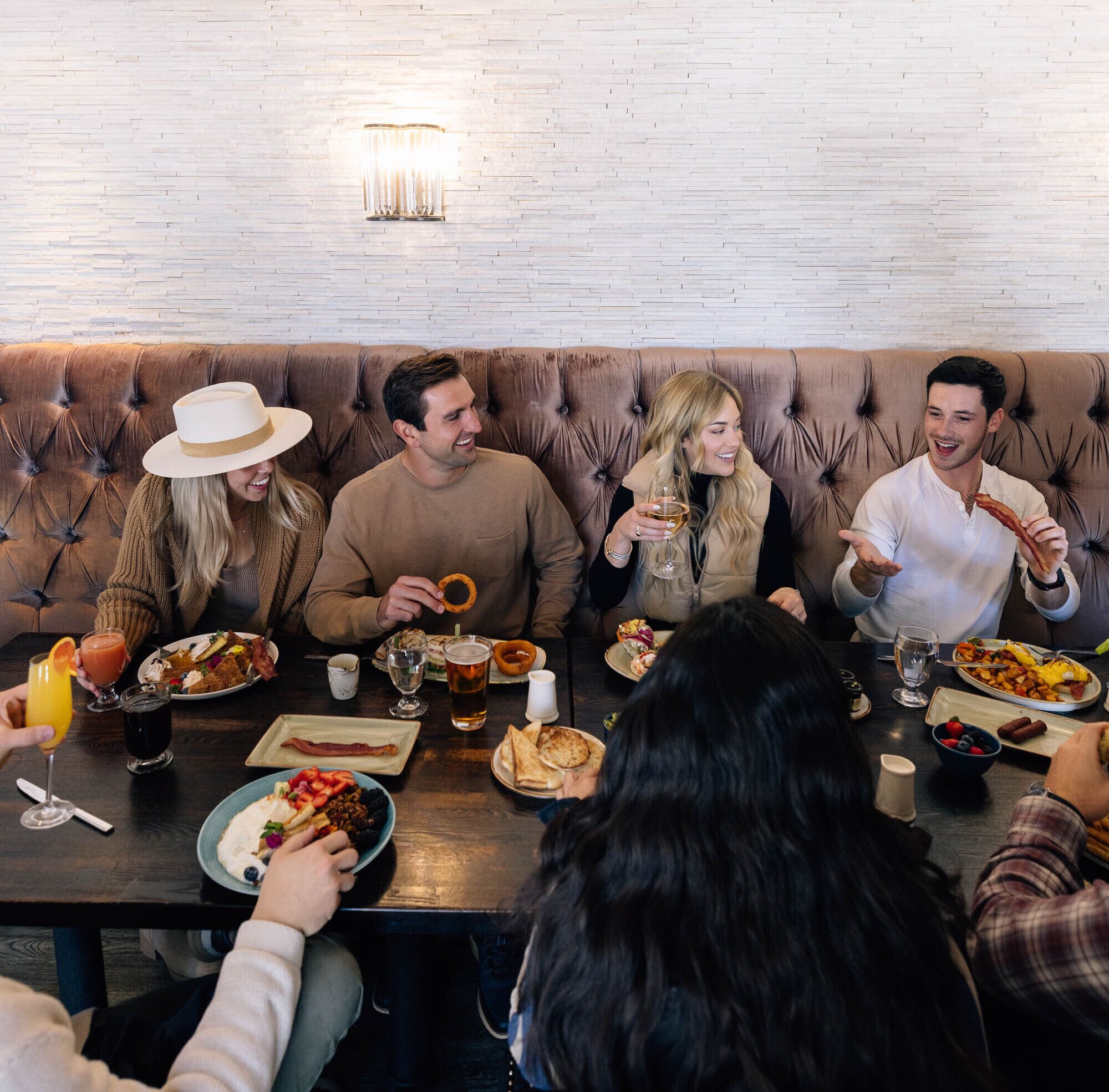 Un groupe de personnes est assis autour d’une table en bois, devant divers plats et boissons, dans une pièce dont un mur est en briques et qui est agrémentée d’un banc en cuir capitonné.
