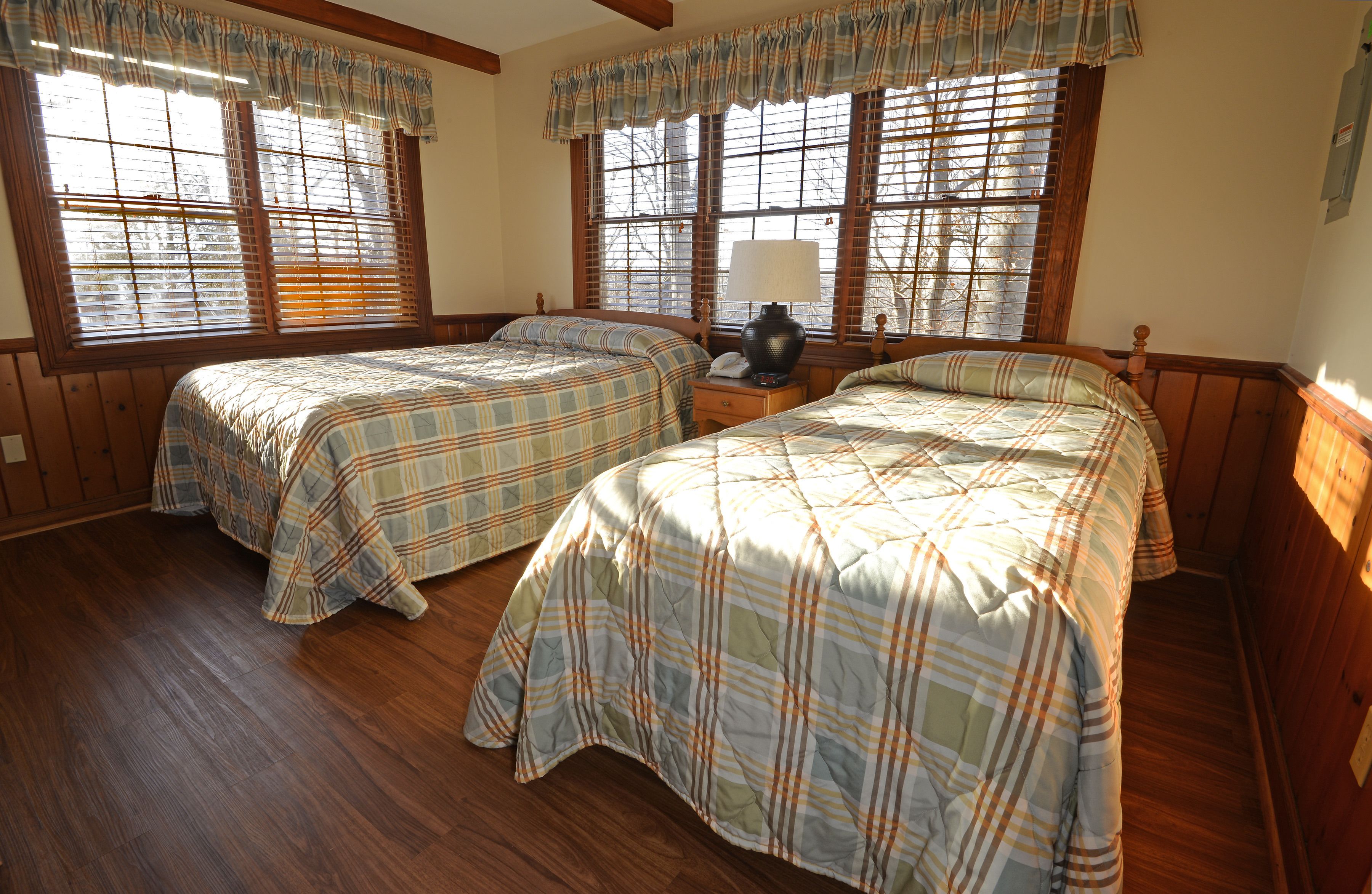 A room with two beds, wooden flooring, and large windows with blinds.