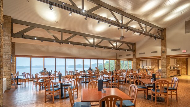 Un vaste espace dédié à la salle à manger, avec des tables et des chaises en bois, de grandes fenêtres offrant une vue sur l’extérieur et un plafond haut avec des poutres apparentes.