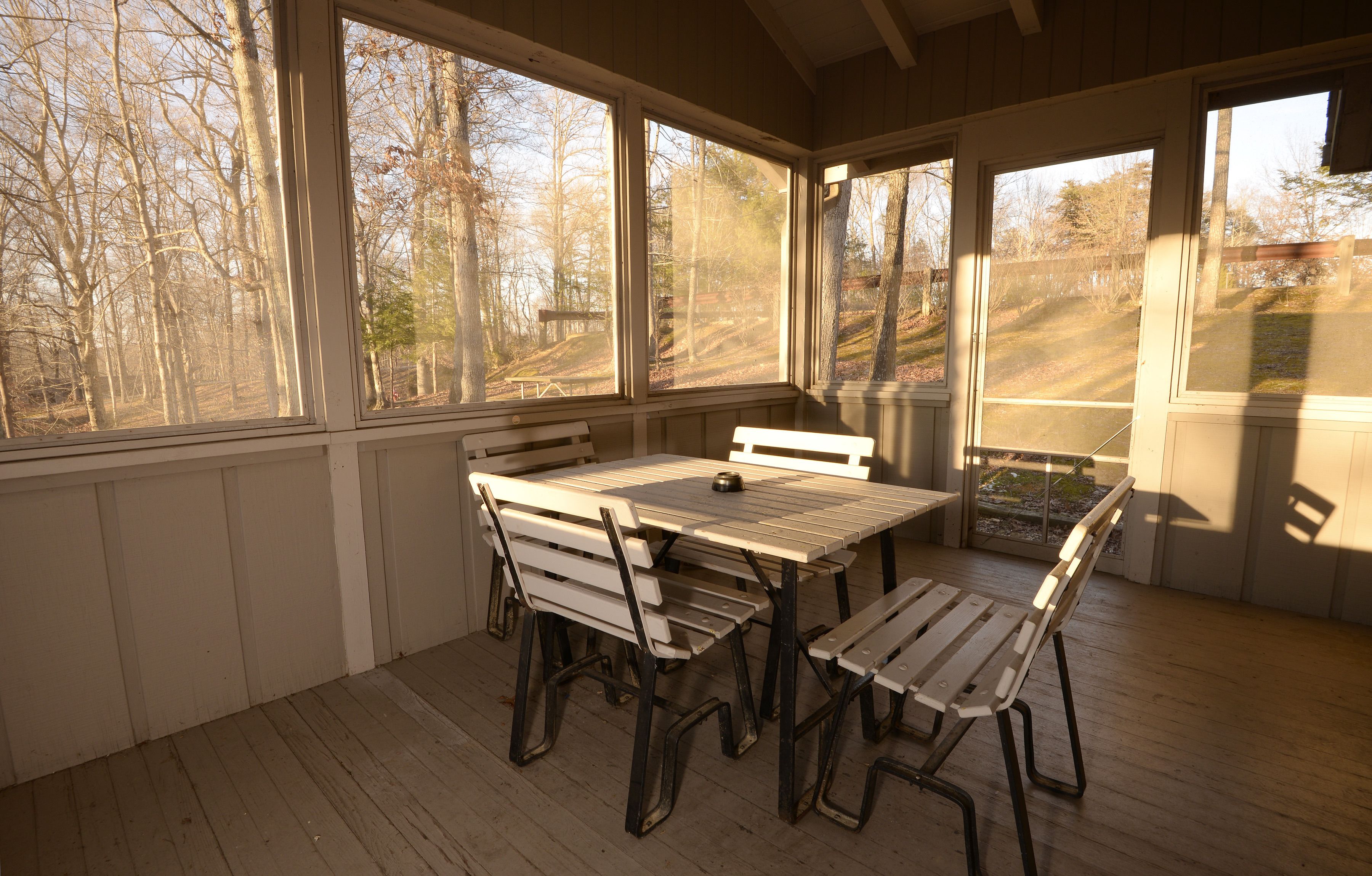 A covered outdoor dining area with a wooden table and benches, large windows, and a view of trees.