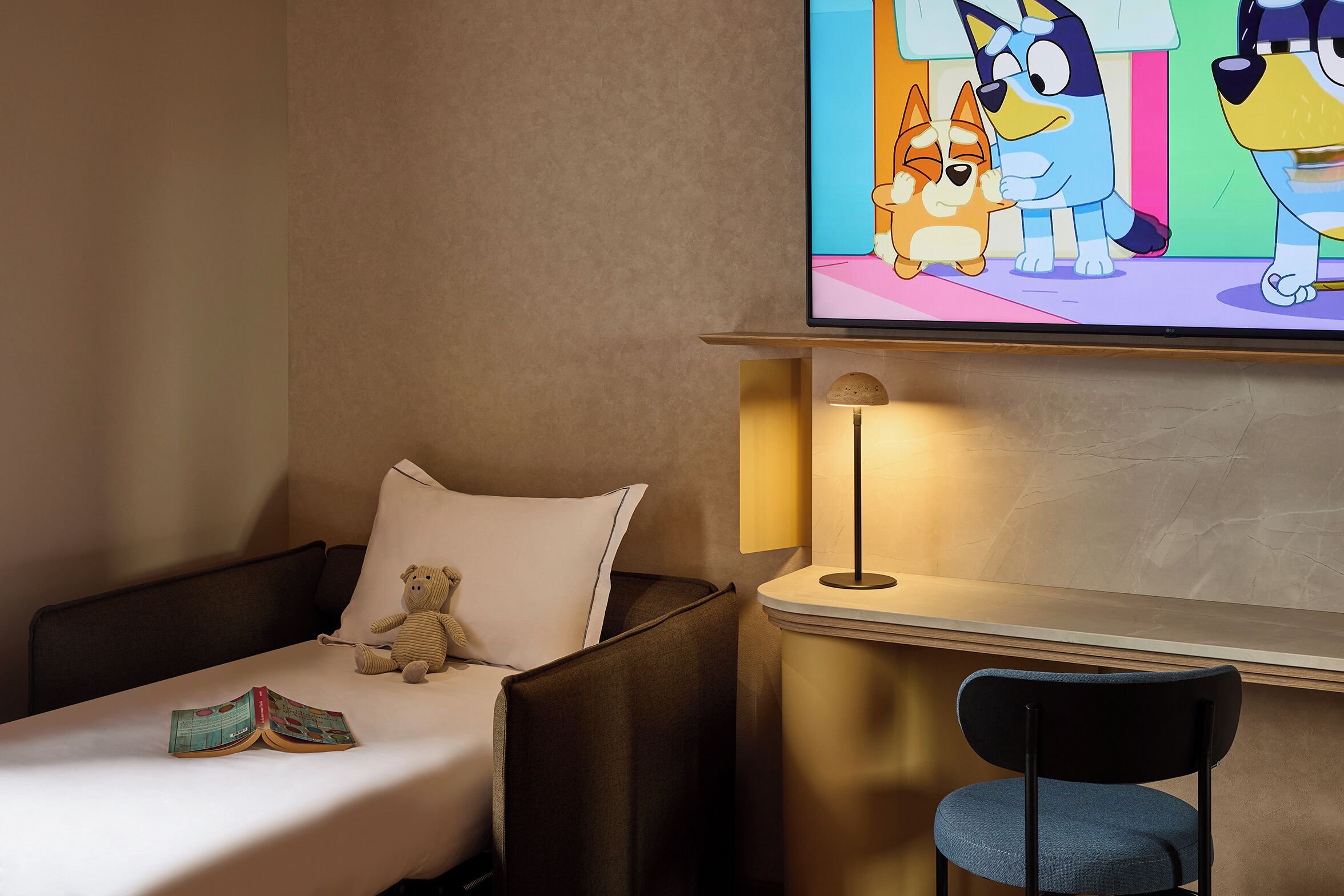 Habitación de hotel con una cama, una silla, una lámpara y un televisor que muestra personajes animados.