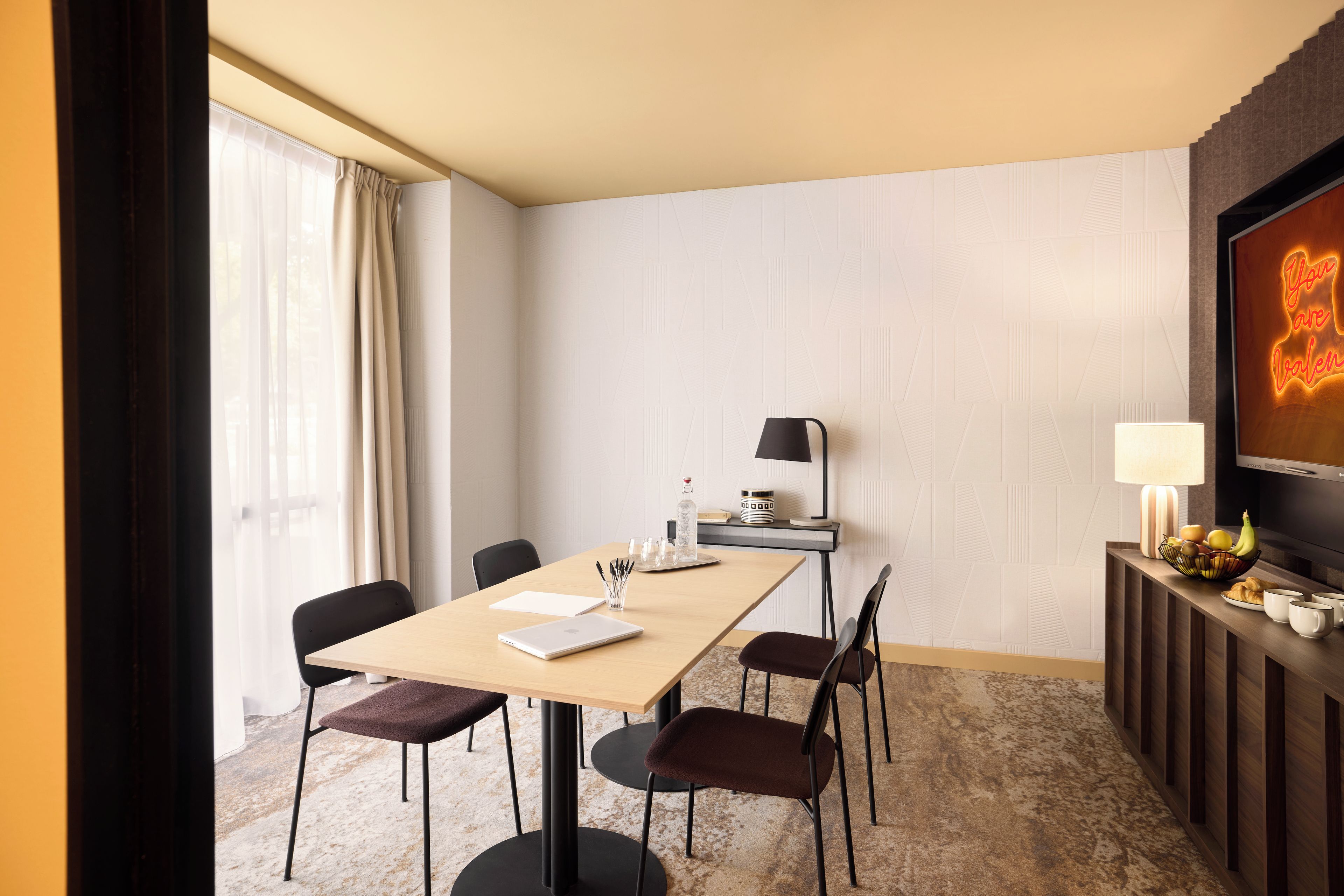 A hotel room with a wooden table, four chairs, a laptop, a lamp, a fruit bowl, and a television.
