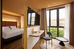 A hotel room with a bed, a desk with a lamp, a chair, and a view of buildings through large windows.