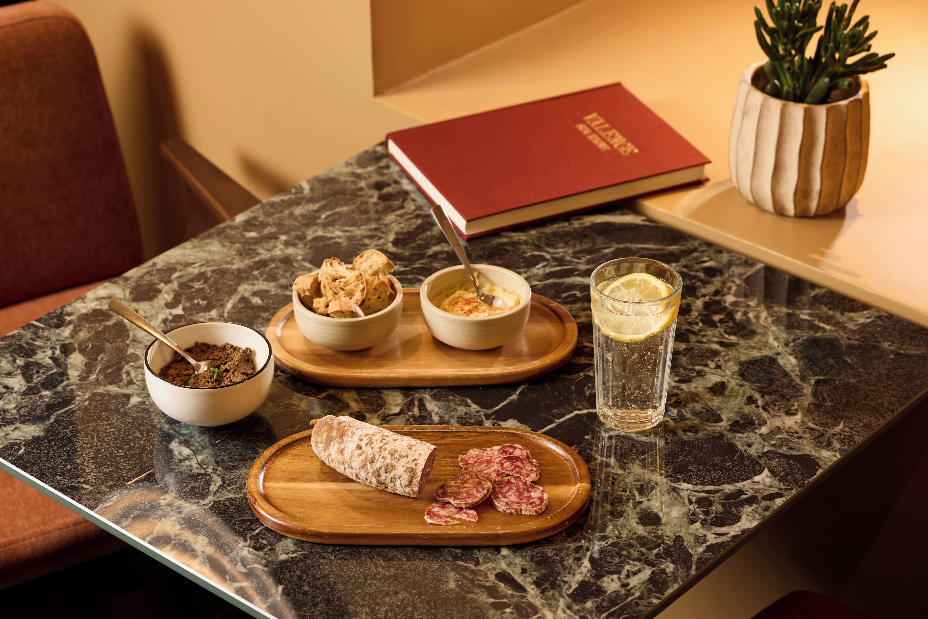 A marble table with a variety of food items including bread, a bowl of what appears to be a spread, a glass of water with a lemon slice, and a book.