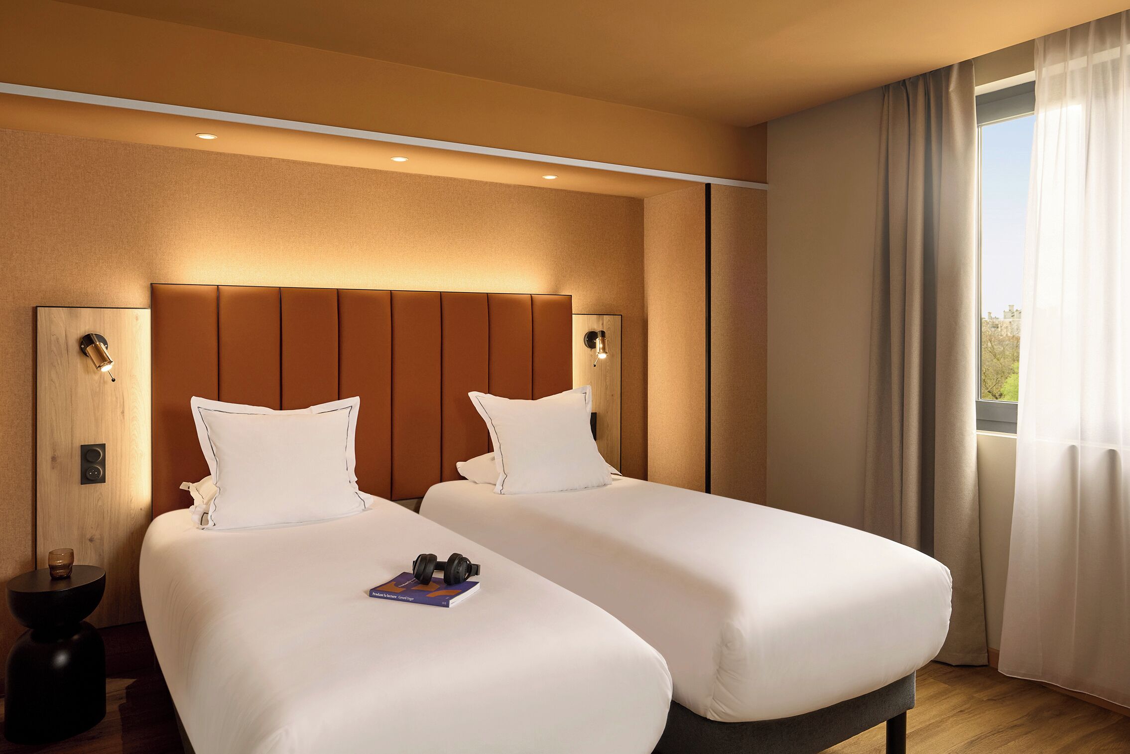 A hotel room with two beds, a bedside table with a book and headphones, a wooden floor, and a window with curtains.