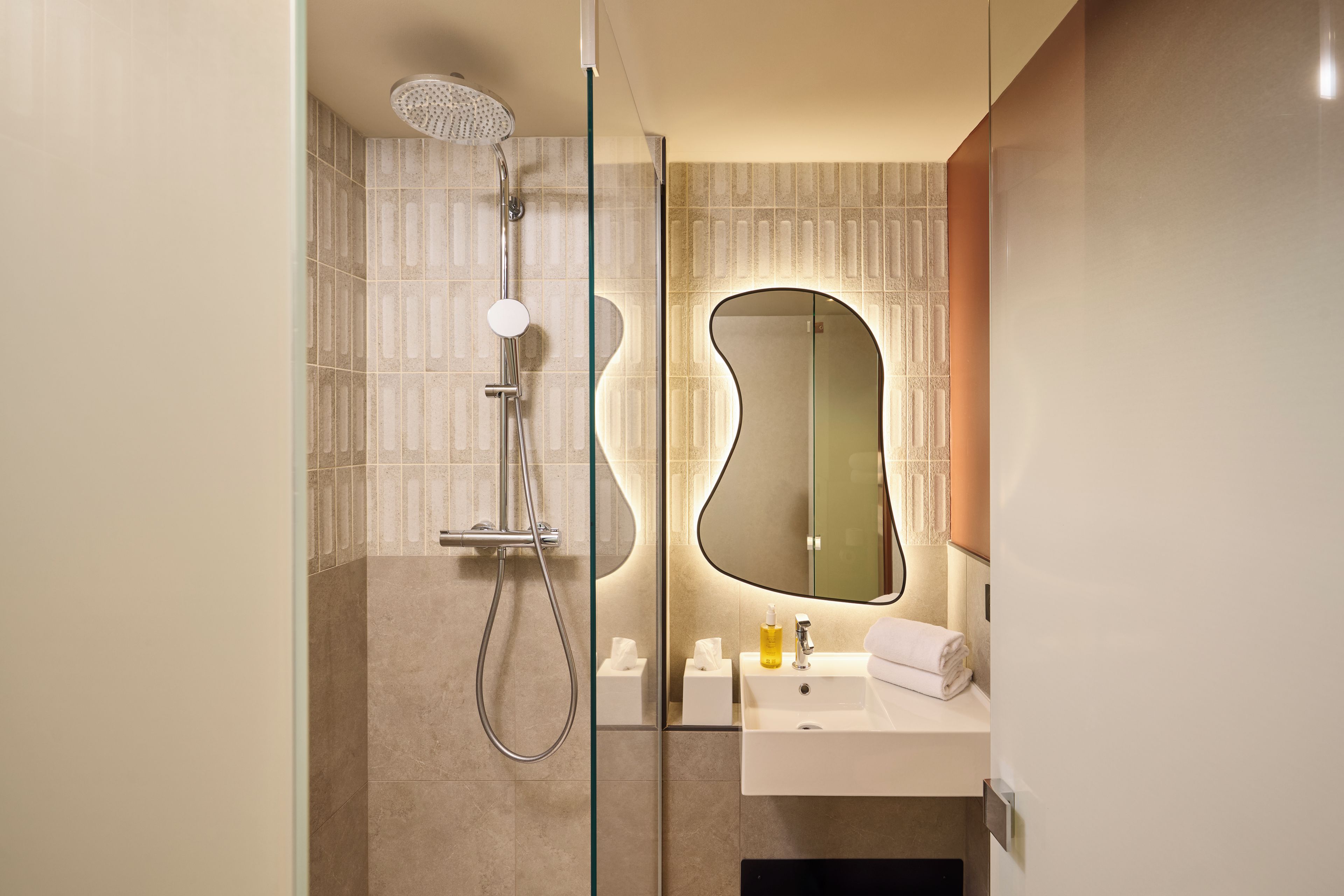Ein modernes Badezimmer mit Duschkabine aus Glas, einem weißen Waschbecken und einem Spiegel mit ungewöhnlicher Form.