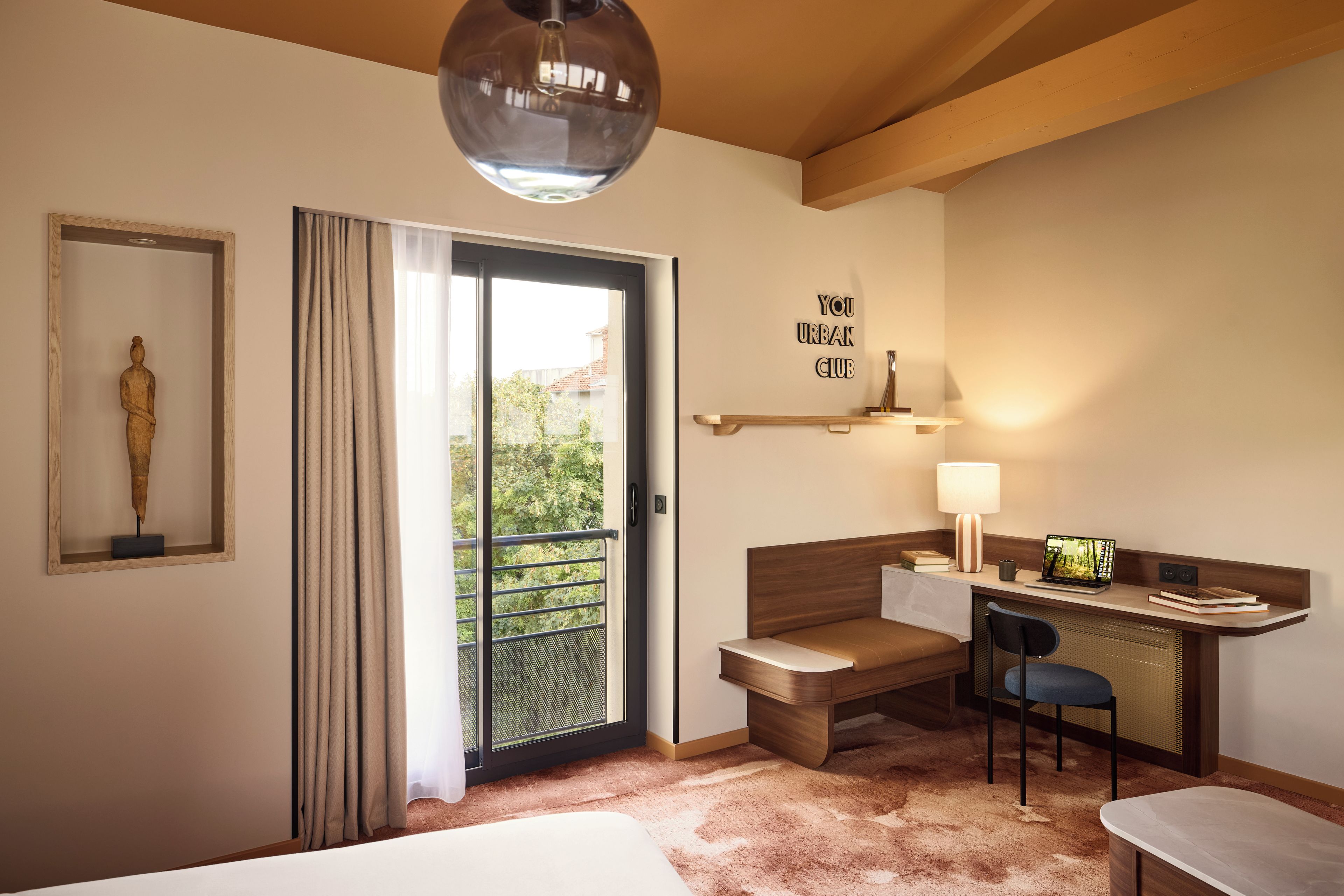 Habitación de hotel con cama, escritorio con silla, lámpara, una pequeña estatua y un cartel en la pared.
