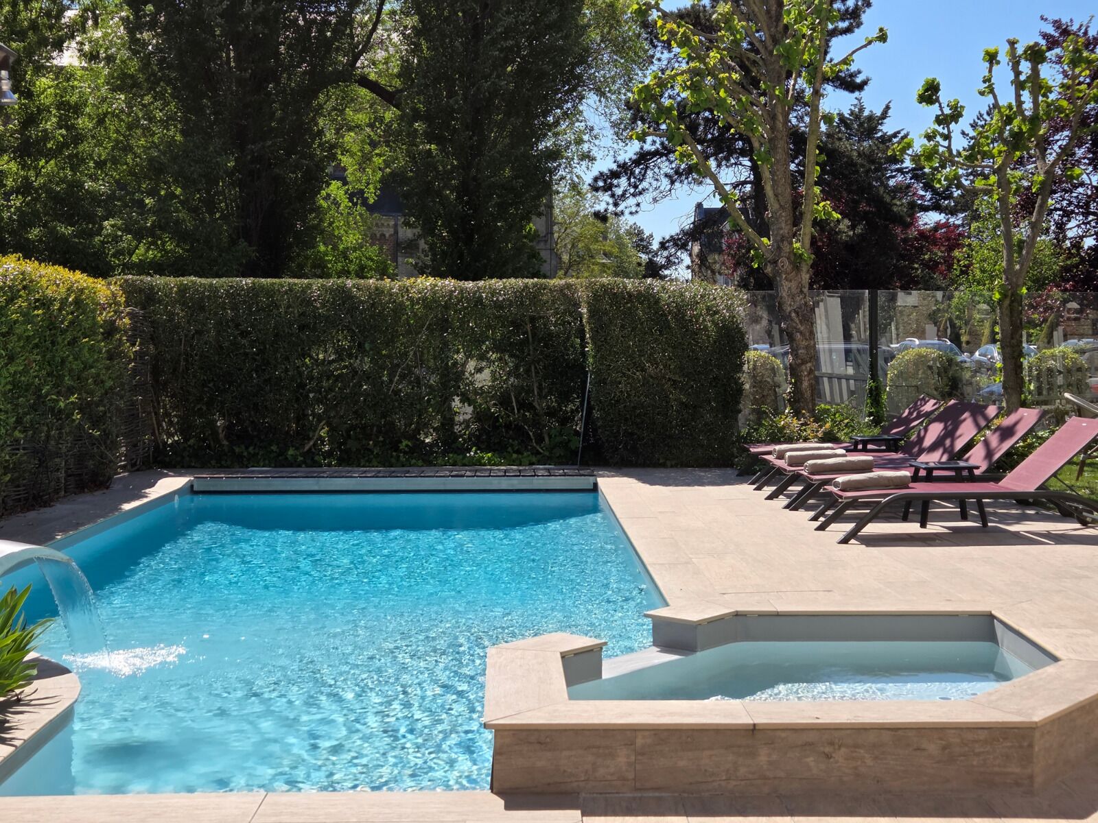 A rectangular swimming pool with a clear blue water, surrounded by a low hedge and a few trees. There are lounge chairs and a hot tub area visible.