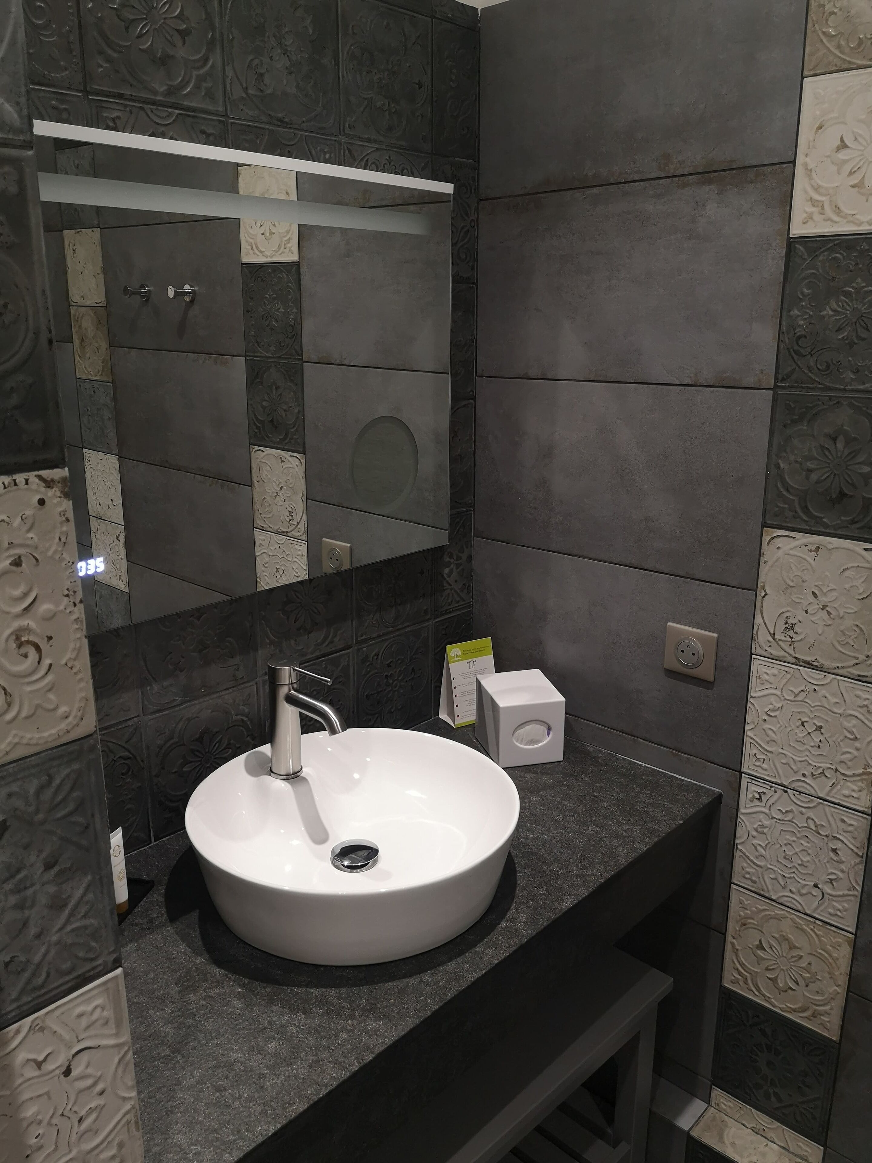 Un baño moderno con un lavamanos blanco, baldosas oscuras y un espejo.