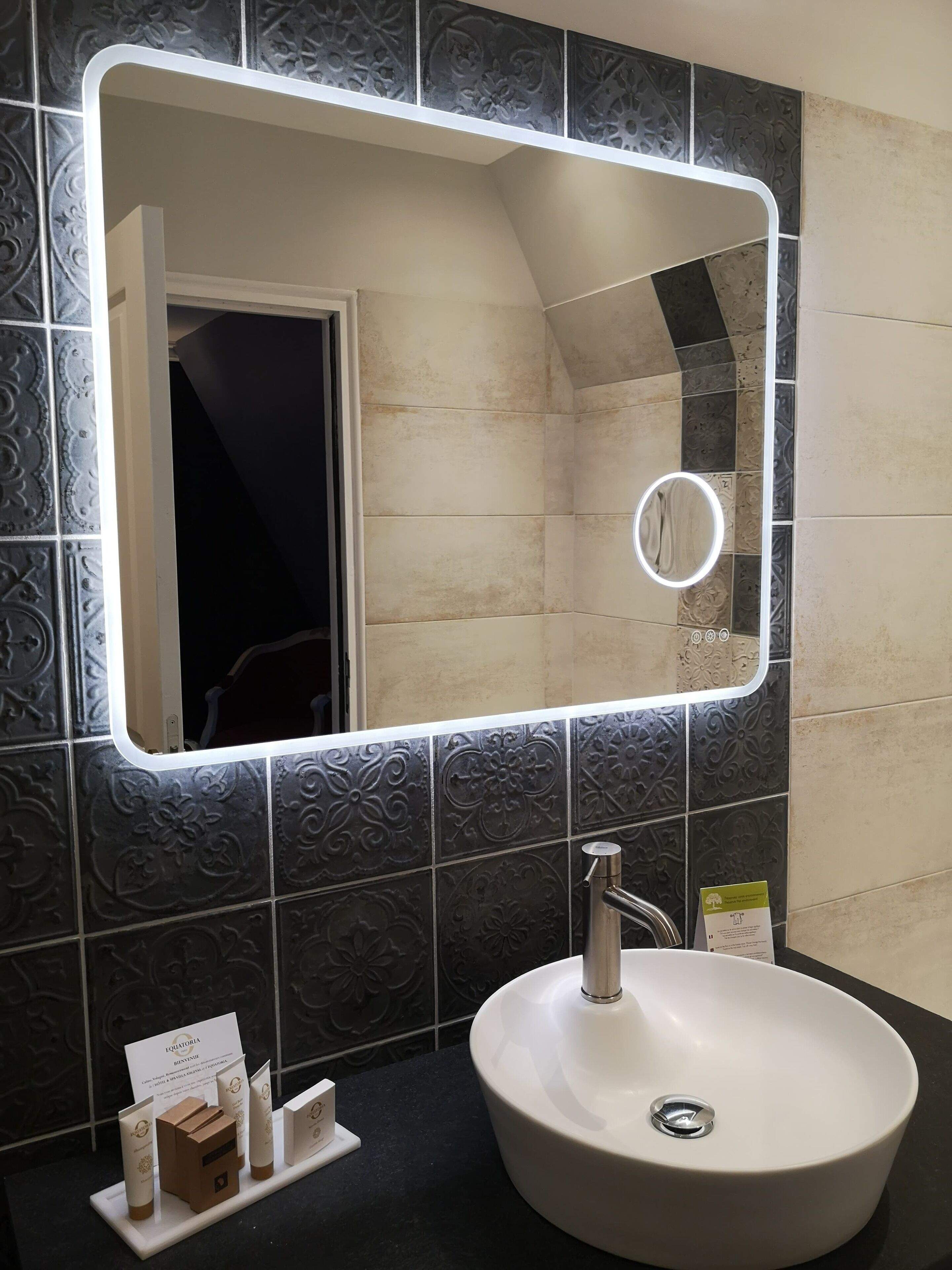 A bathroom with a white sink, a mirror with a light, and toiletries on a shelf.