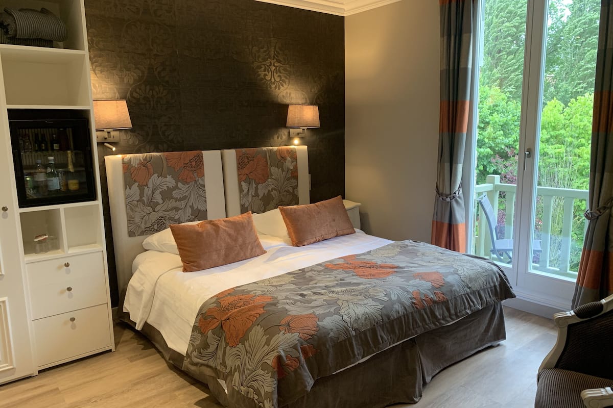 A bedroom with a large bed, a nightstand, a wardrobe, and a window with a view of greenery.