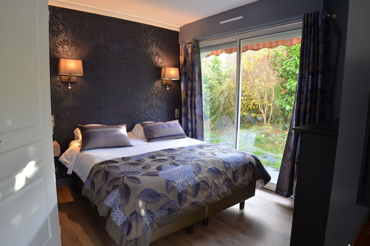 A bedroom with a large bed, two wall-mounted lamps, and a view of greenery through a sliding glass door.