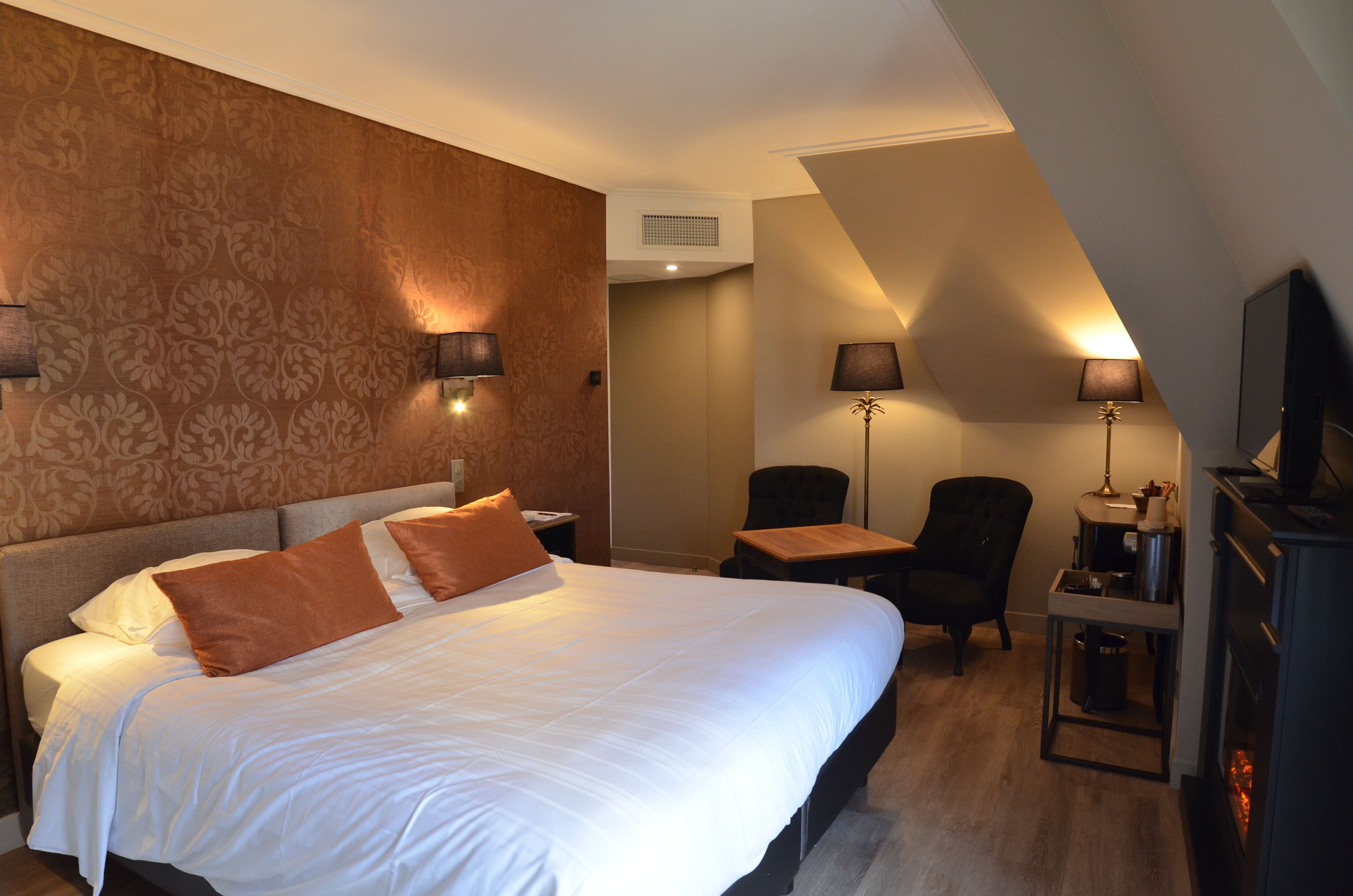 A hotel room with a large bed, two bedside tables, a chair, a small table, a television, and a fireplace.