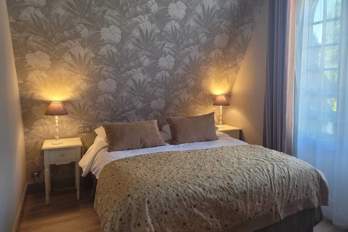 A bedroom with a floral wallpaper, two bedside lamps, a bed with a patterned bedspread, and a window with curtains.
