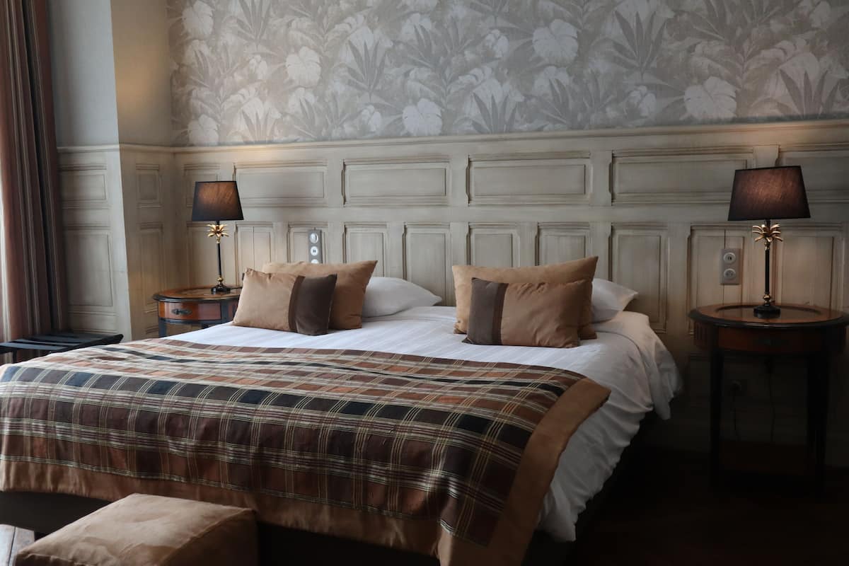 A neatly made bed with a plaid bedspread, two bedside lamps, and a floral wallpaper pattern on the wall.