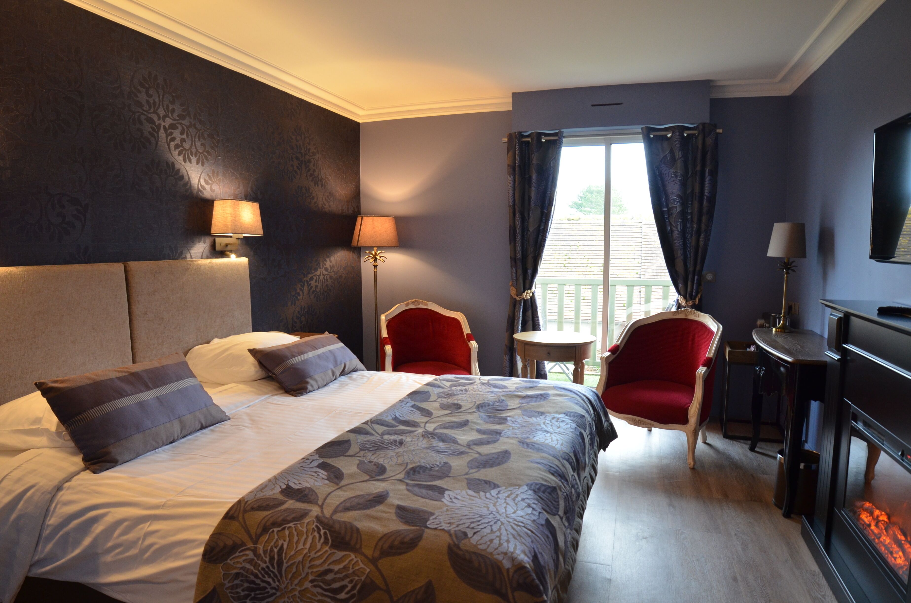 A hotel room with a large bed, two chairs, a small table, a fireplace, and a window with curtains.