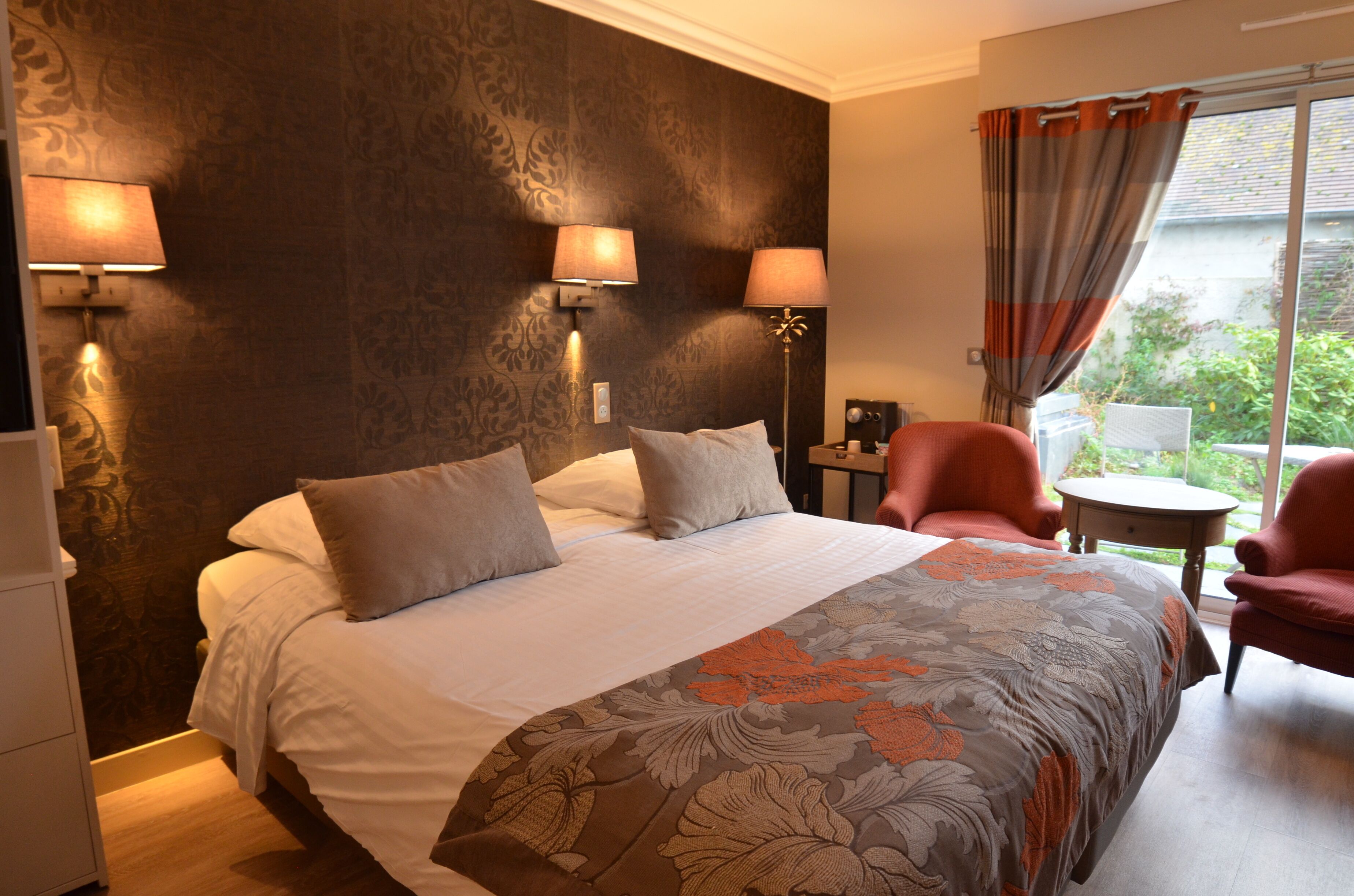 A hotel room with a large bed, two wall-mounted lamps, a chair, a small table, and a view of greenery outside.