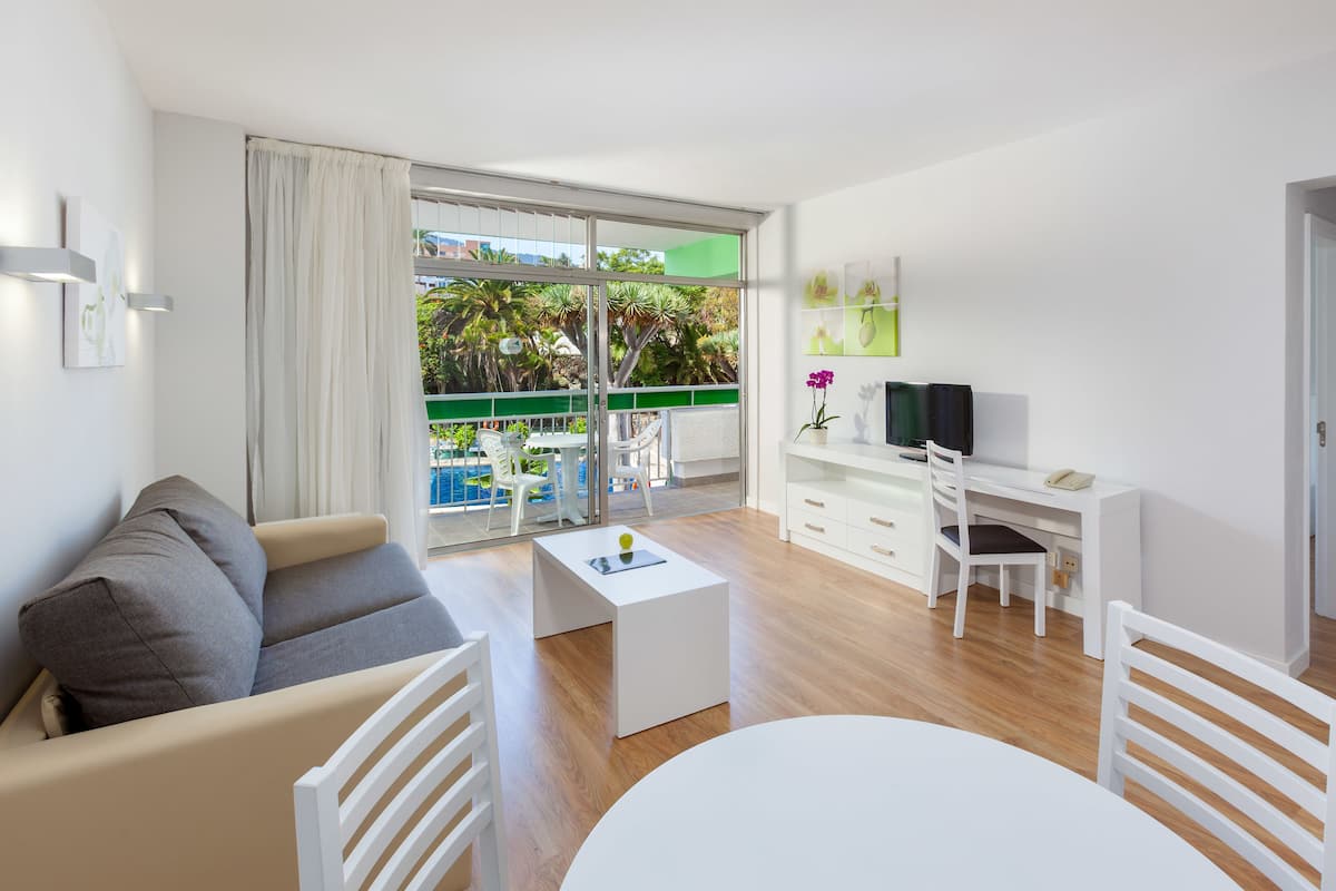 Una sala moderna con un sofá, una mesa de centro y un comedor con mesa y sillas. Se ve un balcón con piscina y vegetación a través de puertas de vidrio.