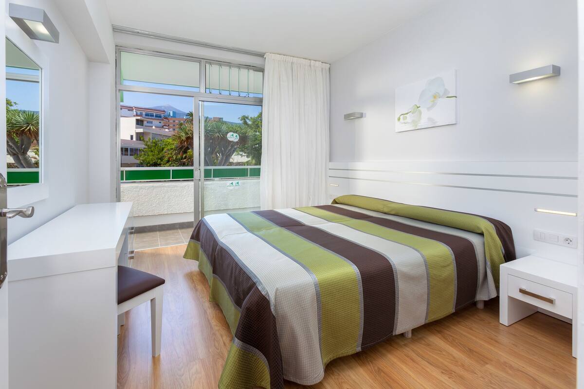 Una habitación de hotel moderna con una cama grande, un escritorio y un balcón con vistas a edificios y zonas verdes.