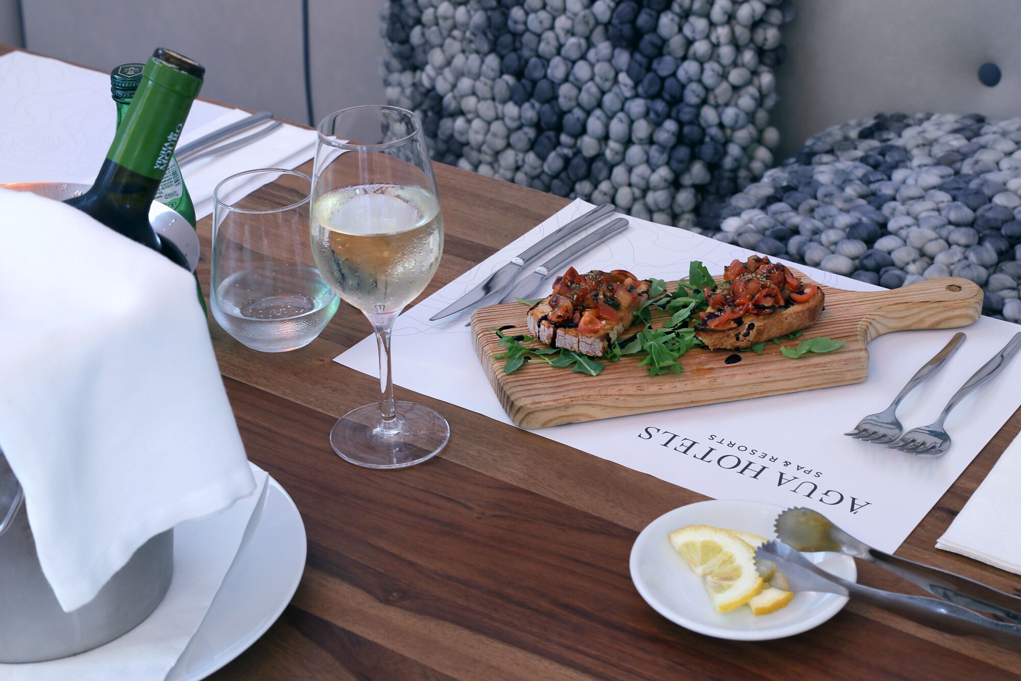 Una mesa de comedor con una tabla de madera, una copa de vino blanco y un plato de comida.