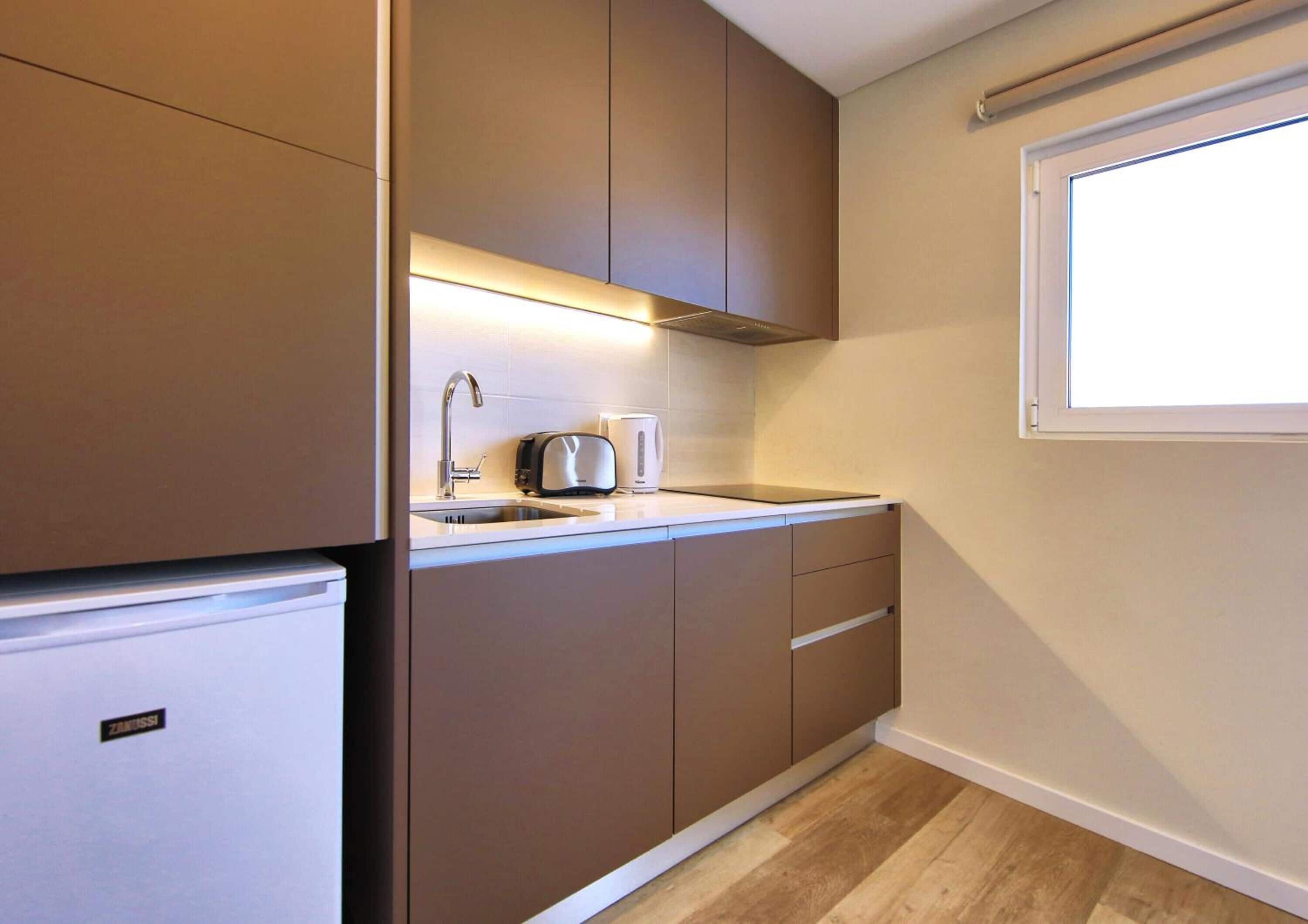 Una cocina compacta con refrigerador incorporado, un lavamanos, un tostador y una tetera.